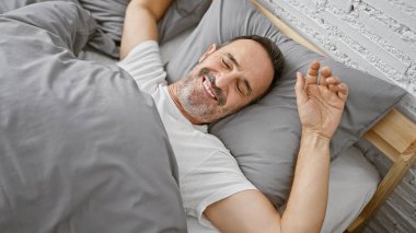 Işıldayan orta yaşlı, gri saçlı, kollarını esneten, rahat yatak odasında uyanan, dinlendirici bir sabahın keyfini çıkaran bir adam.