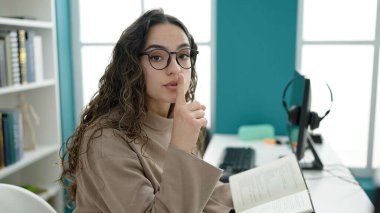 Young beautiful hispanic woman student reading book doing silence gesture at library university