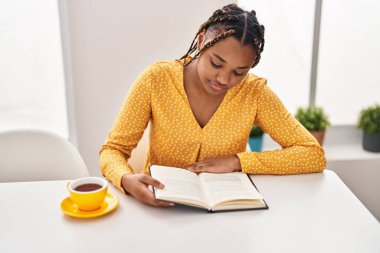 African american woman reading book and drinking coffee sitting on table at home