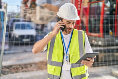 Young hispanic man architect talking on smartphone using touchpad at street