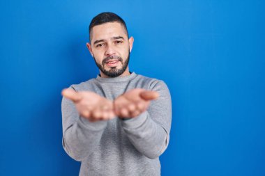 Mavi arka planda dikilen İspanyol adam avuçlarını birbirine uzatarak gülümsüyor ve jest yapıyor. Bekleme ve koruma 