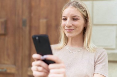 Sokakta akıllı telefon kullanan, kendine güvenen genç sarışın kadın.