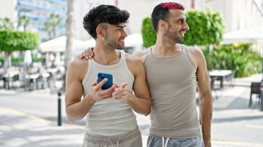 Two men couple smiling confident using smartphone at coffee shop terrace