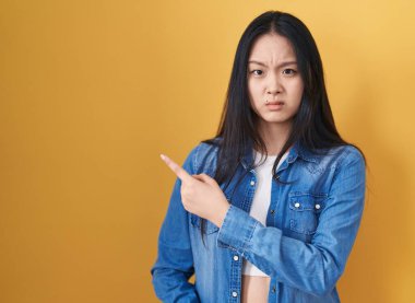 Sarı arka planda duran genç Asyalı kadın endişeli ve endişeli bir ifadeye işaret ediyor. 