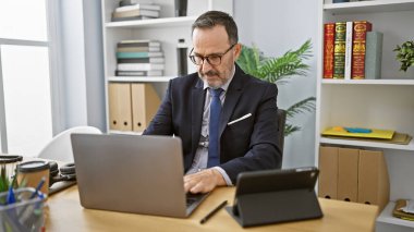 Gri saçlı orta yaşlı bir adam, profesyonel bir iş adamı, ofiste fazla mesai yapıyor, yoğunlaşmış ama rahat, kapalı mekandaki teknolojinin ortasında sakallı ve zarif bir takım elbise.