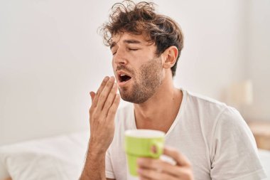 Genç adam bir fincan kahve içiyor yatak odasında esniyor.