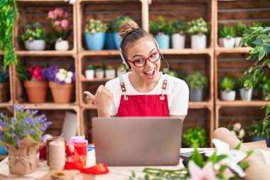 Young hispanic woman working at florist shop doing video call pointing thumb up to the side smiling happy with open mouth 