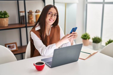 Akıllı telefon ve dizüstü bilgisayar kullanan genç, güzel İspanyol kadın evde masada oturuyor.