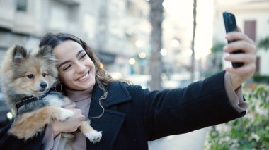 Köpekli genç İspanyol kadın gülümsüyor. Kendine güveni tam. Sokakta akıllı telefondan selfie çekiyor.