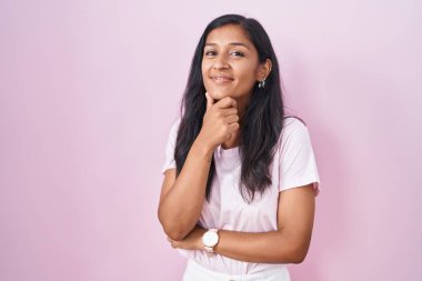 Pembe arka planda duran genç İspanyol kadın kameraya kendinden emin bir şekilde bakıyor kollarını kavuşturmuş ve çenesini kaldırmış gülümsüyor. Olumlu düşünmek. 