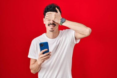 İspanyol adam kırmızı arka plan üzerinde akıllı telefon kullanıyor. Gülümsüyor ve elleri yüzünde, şaşırtmak için gözleri örtüyor. kör kavram. 