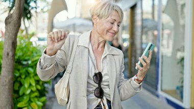 Orta yaşlı sarışın kadın sokakta akıllı telefondan müzik dinliyor.