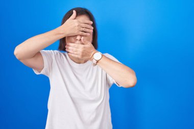 Orta yaşlı İspanyol kadın mavi arka planda dikiliyor gözleri ve ağzı elleriyle kaplıyor, şaşırmış ve şok olmuş. duygu gizleme 