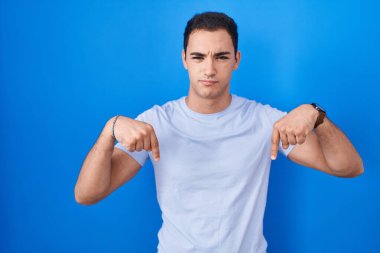 Mavi arka plan üzerinde duran genç İspanyol adam üzgün ve üzgün görünüyor, parmaklarıyla yön gösteriyor, mutsuz ve depresif.. 