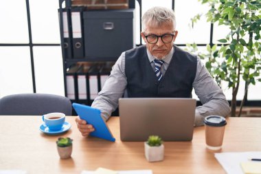 Ofiste dizüstü bilgisayar ve dokunmatik ped kullanan orta yaşlı, kır saçlı bir iş adamı.