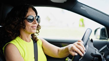 Orta yaşlı İspanyol bir kadın yolda gülümseyen güneş gözlüklü bir araba kullanıyor.
