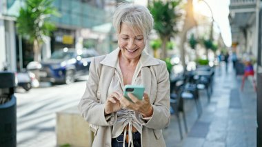 Orta yaşlı sarışın kadın kahve dükkanının terasında akıllı telefon kullanarak gülümsüyor.