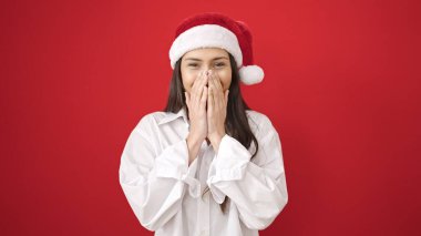 Genç, güzel, İspanyol bir kadın sürpriz bir ifadeyle izole edilmiş kırmızı arka planda noel şapkası takıyor.