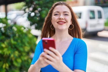Caddede akıllı telefon kullanan, kendine güvenen genç kızıl saçlı kadın.