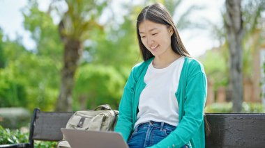 Genç Çinli kadın parkta bankta dizüstü bilgisayar kullanıyor.