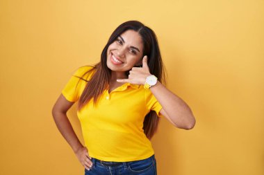 Sarı arka planda duran genç Arap kadın gülümseyerek el ve parmaklarıyla telefon hareketi yapıyor telefonda konuşuyor gibi. iletişim kavramları. 