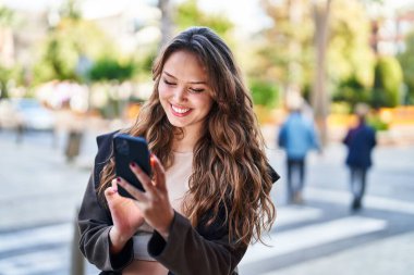 Genç, güzel, İspanyol bir kadın sokakta akıllı telefon kullanarak gülümsüyor.