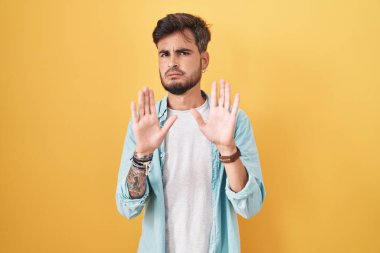 Sarı arka planda duran İspanyol dövmeli genç adam. Avuçlarını hareket ettiriyor. Korkmuş ve iğrenç bir ifadeyle reddediyor. Dur ve yasakla. 