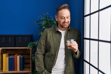 Genç beyaz adam evde dikilip bir bardak viski içiyor.