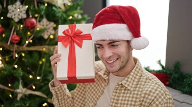 Noel şapkası takmış gülümseyen İspanyol genç adam evde hediyesini dinliyor.