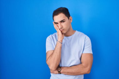Young hispanic man standing over blue background thinking looking tired and bored with depression problems with crossed arms. 
