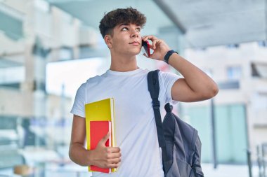 Genç İspanyol öğrenci üniversitede elinde kitaplarla akıllı telefondan konuşuyor.