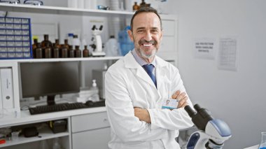 Gülümseyen gri saçlı orta yaşlı bilim adamı kollarını kavuşturuyor, laboratuvarında mikroskopla çalışıyor.