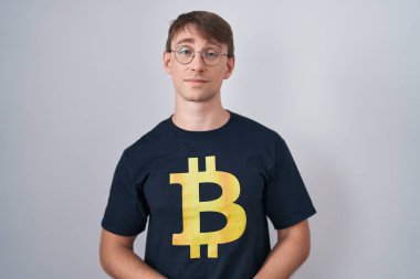 Beyaz, sarışın, bitcoin tişörtlü, elleri çapraz parmaklı, gevşemiş ve neşeli gülümseyen bir adam. Başarı ve iyimserlik 