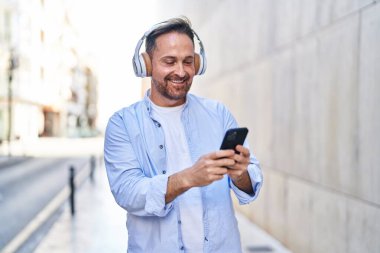 Genç, beyaz bir adam gülerek sokakta müzik dinliyor.