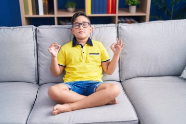 Adorable hispanic boy doing yoga exercise sitting on sofa at home