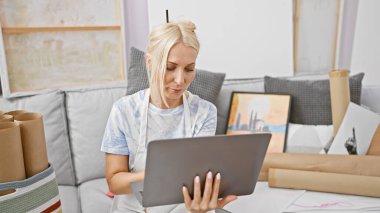 Ciddi genç sarışın sanatçı, konsantre ve odaklanmış, atölye koltuğunda oturuyor, online resim dersi için dizüstü bilgisayar kullanıyor, çizim ve resim dünyasına dalmış.