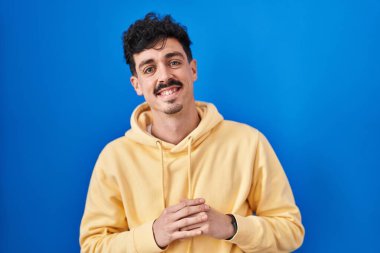 Mavi arka planda dikilen İspanyol adam ellerini birleştirip gülümseyerek gevşemiş ve neşeli olmasını diledi. Başarı ve iyimserlik 