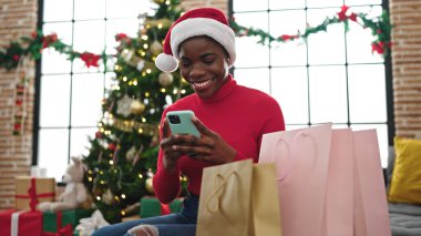 Afro-Amerikalı kadın akıllı telefon kullanıyor. Kanepede oturuyor ve evde Noel hediyesi çantalarıyla oturuyor.