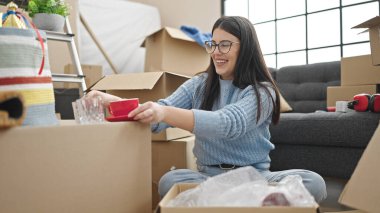 Young hispanic woman smiling confident unpacking cardboard box at new home