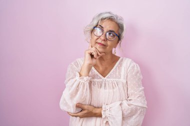 Pembe arka planda beyaz saçlı orta yaşlı bir kadın, elleri çenesinde soru sorarken, düşünceli bir ifade takınıyor. Düşünceli bir yüzle gülümsüyor. şüphe kavramı. 
