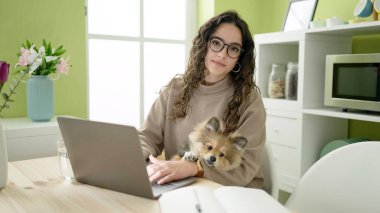 Yemek odasında laptopunu kullanan köpekli genç İspanyol kadın.