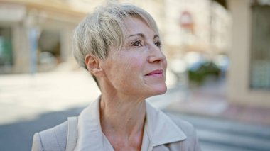 Orta yaşlı sarışın kadın sokakta ciddi bir ifadeyle yan tarafa bakıyor.