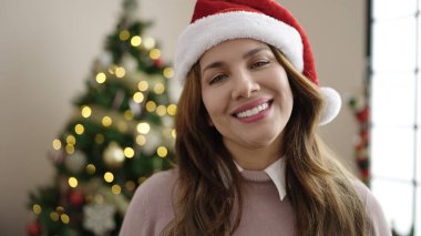 Genç, güzel, İspanyol bir kadın. Gülümsüyor. Noel ağacının yanında duruyor.