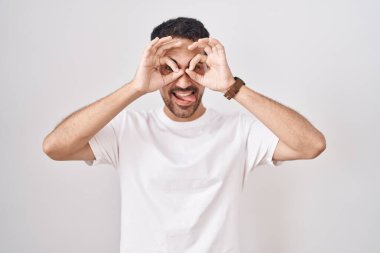 Beyaz arka planda duran yakışıklı İspanyol adam dürbün gibi jest yapıyor. Dürbün gibi dilini çıkarıyor, gözler parmaklarından bakıyor. çılgın ifade. 