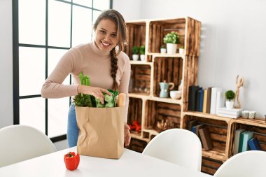 Genç, güzel, İspanyol bir kadın gülümsüyor. Elinde alışveriş torbası var.