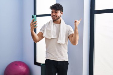 Sakallı İspanyol bir adam spor salonunda akıllı telefonuyla video görüşmesi yapıyor. Gülümsüyor, mutlu bir şekilde elini ve parmağını yana uzatıyordu. 