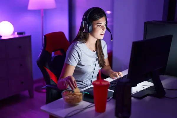 Young beautiful hispanic woman streamer playing video game eating chips potatoes at gaming room