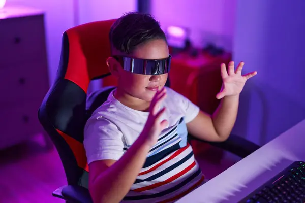 Adorable hispanic boy streamer playing video game using virtual reality glasses at gaming room