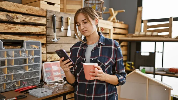 Young blonde woman carpenter using smartphone drinking coffee at carpentry