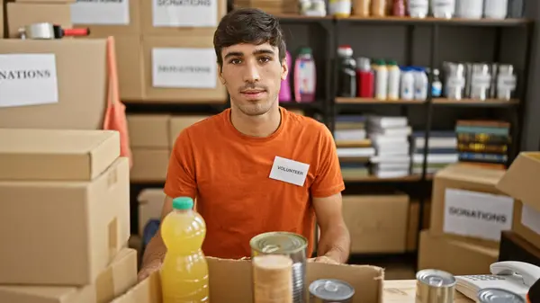 Devoted young hispanic man actively volunteering at a charity center, sitting at the table with a serious face, working diligently in a room full of donations